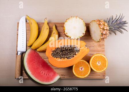 Ein Schneidebrett mit geschnittenem Obst und einem Messer, eine Papaya, eine Wassermelone, eine Ananas, Orangen und Bananen Stockfoto