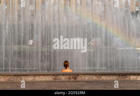 Kopf und Schultern einer Frau, Rückansicht, gegen den sprenkelnden Brunnen, mit Regenbogen, an einem heißen Sommertag Stockfoto