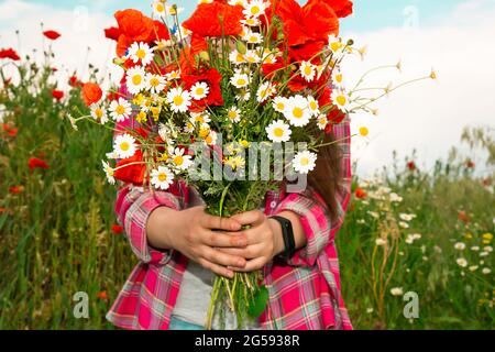 Das Mädchen steht vor dem Hintergrund wilder Blumen und hält einen Strauß Mohnblumen und Gänseblümchen in den Händen. Ein Mädchen auf dem Feld sammelte einen Strauß wilder Blumen. Stockfoto