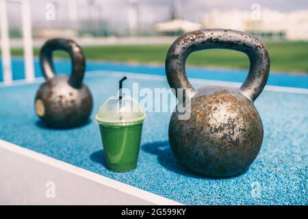 Gesundheit und Fitness Green Smoothie Detox Drink im Fitnessstudio mit Kettlebells Gewichten im Outdoor Training Fitness Center. Plastikbecher Gemüsesaft Stockfoto