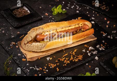 Hot Dog mit Gurken, getrockneten Zwiebeln und Mustar auf schwarzem Schiefer Hintergrund, Stillleben Stockfoto