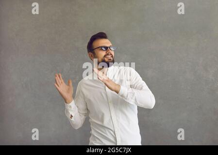 Fröhlicher junger Mann in einer zermollten Lebensbrille, der auf grauem Hintergrund zu Musik tanzt Stockfoto