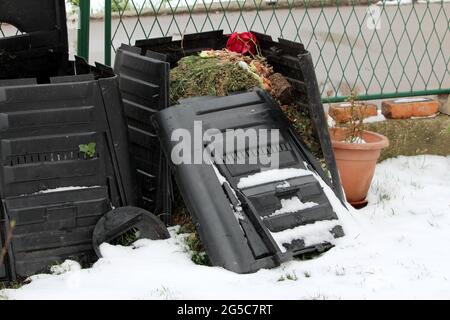 Zerbrochene alte zerstörte fast vollständige schwarze Plastikkompostbehälter neben Blumentopf und Metallzaun im Hintergrund mit frisch gefallener Schnee bedeckt Stockfoto