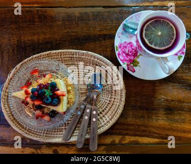 Gemischter Beerenkochkuchen auf einer Glasplatte über einem kleinen Rattan-Tablett zusammen mit einer Tasse Zitronentee im Vintage-Becher-Set auf einem Holztisch im gemütlichen Café. Stockfoto