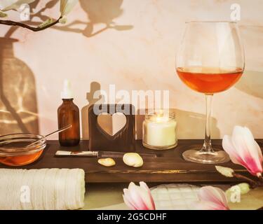 Home Spa Entspannen. Umweltfreundliches Badezimmer mit Spa-Accessoires, Serum, Kerze und einem Glas Roséwein auf einem Holzbrett mit geschnitztem Herz Stockfoto