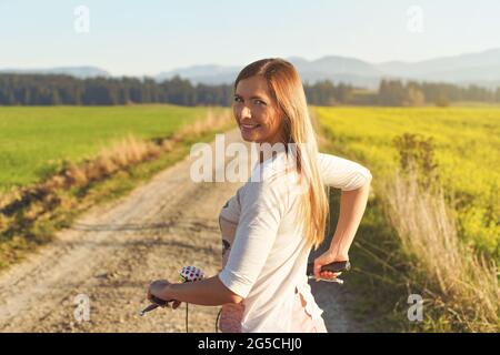 Junge Frau, die neben ihrem Fahrrad auf einer staubigen Landstraße steht und über die Schulter zurückblickt Stockfoto