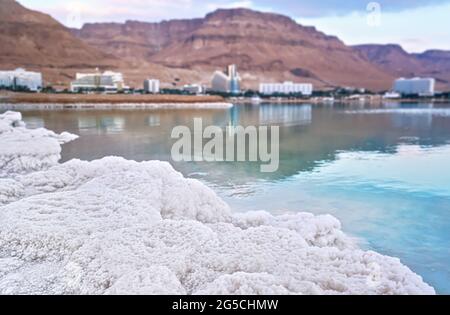 Sand komplett mit kristallinem Salz bedeckt, im Hintergrund verschwommen Hotels und Resorts an der israelischen Küste des Toten Meeres Stockfoto