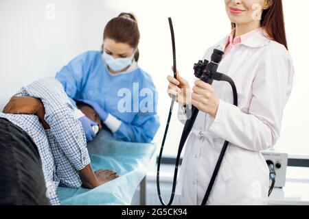 Nahaufnahme einer jungen Ärztin mit Endoskop. Afroamerikanischer Patient auf der Couch liegend und eine Krankenschwester, die ihn auf die Gastroendoskopie vorbereitet. Stockfoto
