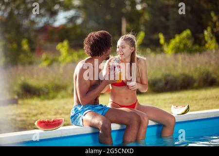 Junges multiethnisches Paar, das am Rand des Pools sitzt, Säfte trinkt, lächelt, lacht und sich gegenseitig anschaut Stockfoto