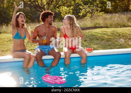 afroamerikanischer junger Mann, der am Rand des Pools mit zwei kaukasischen Weibchen im Freien in der Natur sitzt Stockfoto