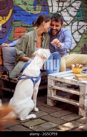 Junges Paar, das Spaß mit seinem Hund im Café im Freien hat, sich gegenseitig kuschelt und das Tier labrador Retriever füttert. Stockfoto