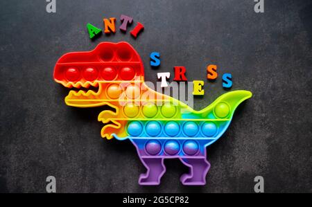 Toy Pop it Dinosaurier Regenbogenfarben auf schwarzem Hintergrund mit bunten Buchstaben und der Aufschrift-Anti-Stress auf dem Rücken. Stockfoto