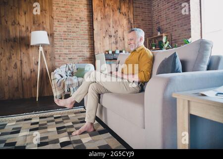 Ganzkörper-Profil Porträt von fröhlich gealterten Mann sitzen auf Sofa-Look Laptop toothy Lächeln tippen Massage nach Hause drinnen Stockfoto