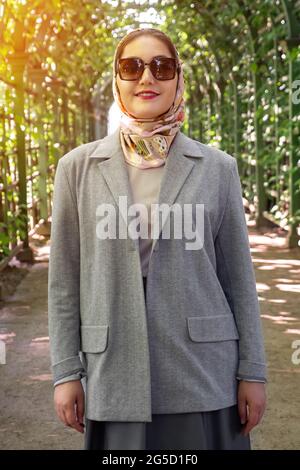 Lächelnde muslimische Frau in grauem Anzug und Sonnenbrille im Park, Nahaufnahme Stockfoto
