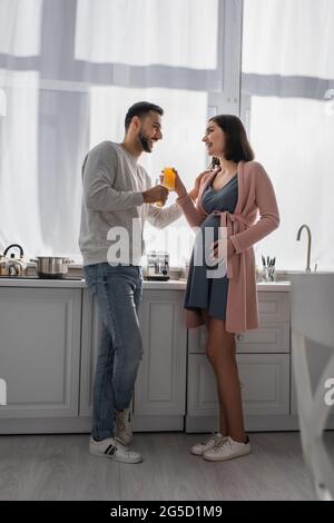 Glücklicher junger Mann und Schwangere stehen mit Orangensaft in der Küche Stockfoto