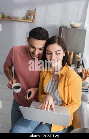Lächelnder junger Mann, der mit Handy und Kaffeetasse in der Nähe einer Freundin steht, die zu Hause mit einem Laptop sitzt Stockfoto