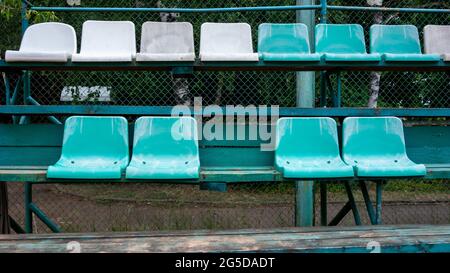 Leere 2 Reihen von einfachen grünen, weißen Retro-Plastikstühlen, die an Holztribünen befestigt sind, um ein Sportspiel zu beobachten. Eine fehlt. Stockfoto