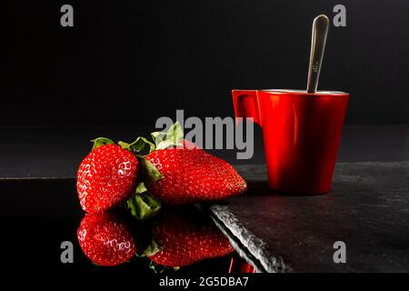 Zwei frische rote Erdbeeren spiegeln sich auf einem Spiegel und eine rote Tasse auf einem Schieferboden.das Foto ist ein Stillleben, das mit künstlichem Licht und horiz aufgenommen wurde Stockfoto