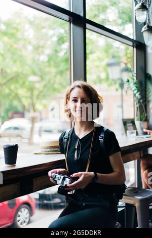 Die junge Hipster-Frau hält eine Retro-Kamera in den Händen und schaut in einem alten Café auf die Kamera Stockfoto