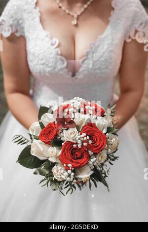 Eine Braut in einem Hochzeitskleid hält einen dekorativen Brautstrauß aus roten und weißen Rosen in ihren Händen, Nahaufnahme. Stockfoto