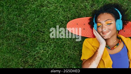 Mixed Race Frau mit Vitiligo trägt blaue Kopfhörer hört Musik auf dem Gras in der Nähe von Skateboard liegen und träumt mit geschlossenen Augen Stockfoto