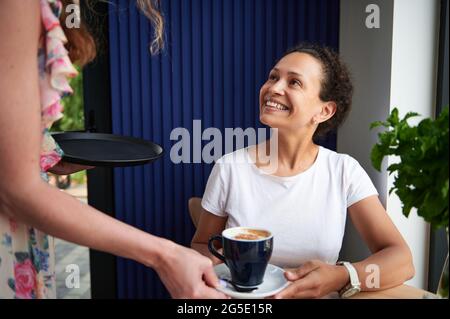 Charmante Brünette Frau sitzt am Tisch im Café und lächelt Kellnerin servieren und bringen eine Tasse mit Cappuccino . Stockfoto