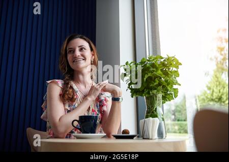 Eine attraktive Frau genießt eine Kaffeepause, während sie in einem Café an einem Tisch am Fenster sitzt Stockfoto