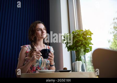 Schöne Frau mit einer Tasse Kaffee in den Händen, die am Tisch neben einem Fenster im Café sitzt. Kaffeepause. Stockfoto