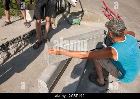 Der Prozess der Verlegung der Bürgersteig-Bordsteinkante. Die Arbeiter messen ein Stück Beton, bevor sie mit einer Kreissäge schneiden. Stockfoto