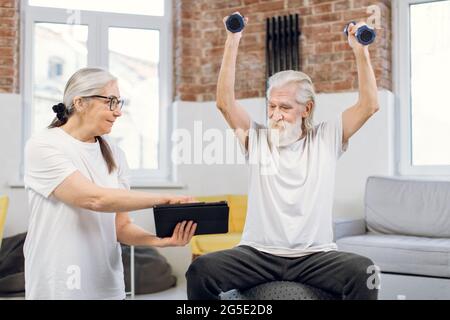 Älterer Mann, der mit Kurzhanteln am Fitball trainiert und sich Online-Tutorials auf einem digitalen Tablet ansieht, das ihre charmante Frau in den Händen hält. Paar mit modernen Gadgets für das Training zu Hause. Stockfoto