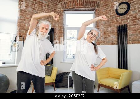 Zwei Rentner, die sich am Morgen zu Hause sportlich betätigen, machen zu Hause Sportübungen. Positives Seniorenpaar, das sich in Innenräumen den Körper streckt. Stockfoto