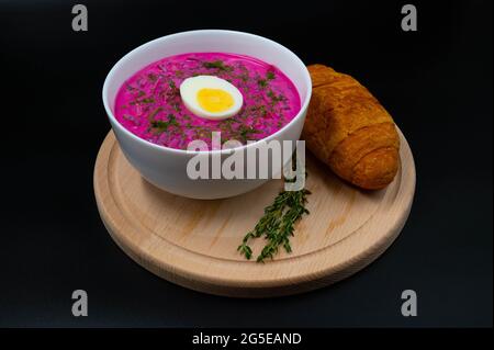 Sommerliche kalte Rote-Beete-Suppe (choldnik) mit gekochtem Ei, serviert in einer Schüssel auf einem Holzbrett mit einem Croissant. Isolierter schwarzer Hintergrund. Stockfoto