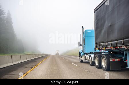 Blauer großer Langstrecken-Sattelschlepper, der kommerzielle Fracht in einem trockenen Transporter transportiert, der auf einer einbahnigen Autobahnstraße mit Wald auf dem hil läuft Stockfoto