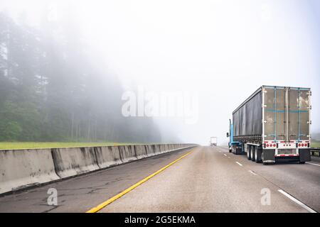 Blauer großer Langstrecken-Sattelschlepper, der kommerzielle Fracht in einem trockenen Transporter transportiert, der auf einer einbahnigen Autobahnstraße mit Wald auf dem hil läuft Stockfoto