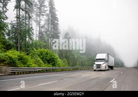 Weißer großer Langstrecken-Sattelschlepper, der kommerzielle Fracht in einem trockenen Kleintransporter transportiert, der auf einer einbahnigen Autobahnstraße mit Wald auf der Hi läuft Stockfoto