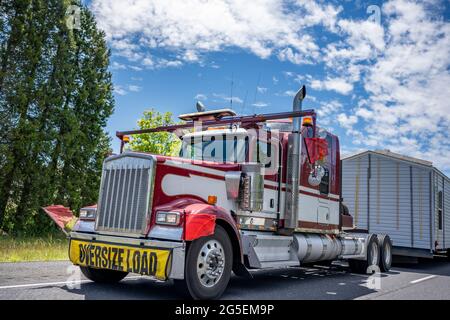 Speziell ausgestattete American Idol Red Classic Big Rig Semi-Truck mit Chromteilen und Übergröße Lastschild auf der Vorderseite transportiert Manufakturhaus Stockfoto