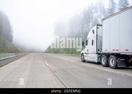 Weißer großer Langstrecken-Sattelschlepper, der kommerzielle Fracht in einem trockenen Kleintransporter transportiert, der auf einer einbahnigen Autobahnstraße mit Wald auf der Hi läuft Stockfoto