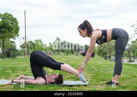 Weibliche Personal Trainer hilft jungen Mann in einem Park, seinen Rücken und Beine zu strecken. Dehnübungen auf einer Matte draußen. Stockfoto
