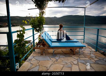 Frau in legerer Kleidung sitzt auf einem Holzdeckstuhl auf einer Terrasse mit Blick auf den See bei Sonnenuntergang, Lin, Ohrid-See, Korca-Region, Albanien Stockfoto