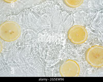 Zitronenscheiben in sauberem, transparentem Wasser auf weißem Hintergrund mit Kopierfläche. Wasser spritzt im Sonnenlicht auf die Wasseroberfläche. Draufsicht oder Flatlay. Sommer, Urlaub, gesunde Ernährung Konzept Stockfoto