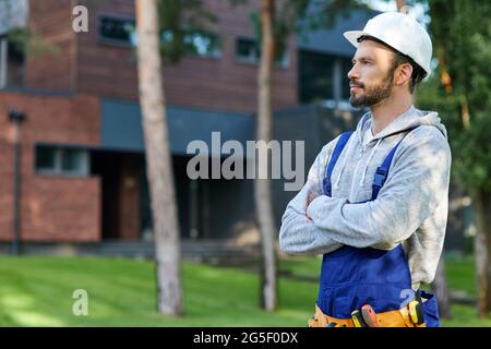 Junger bärtiger Ingenieur mit hartem Hut schaut weg und posiert im Freien, während er an der Hütte arbeitet Stockfoto