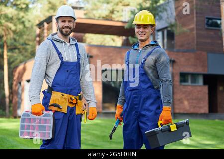Verlassen Sie sich auf uns. Zwei gutaussehende junge Ingenieure in Hartmützen lächeln vor der Kamera und posieren im Freien mit Werkzeugkiste und Koffer mit Werkzeugen Stockfoto