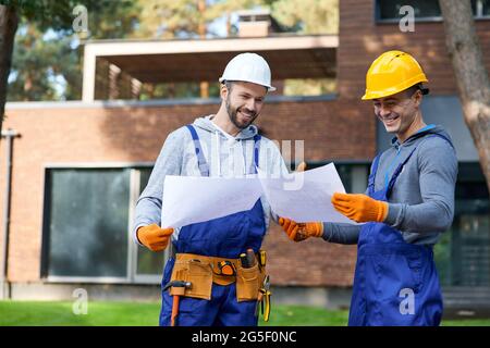 Entscheidungsfindung. Zwei positive Bauherren lächeln, während sie draußen mit einem offenen Plan stehen, der das halb fertiggestellte Cottage House diskutiert Stockfoto