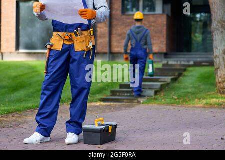 Ausgeschnittene Aufnahme eines männlichen Baumeisters in blauen Overalls, der im Freien mit offenem Bauplan stand, während er am Bau eines Ferienhauses arbeitete Stockfoto