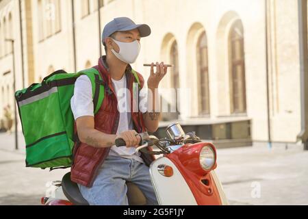 Kontaktlose Lieferung während der Quarantäne. Junger asiatischer männlicher Kurier mit Thermosack, der Gesichtsschutzmaske trägt und mit dem Mobiltelefon spricht Stockfoto