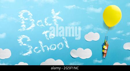 Gelber Ballon, der einen Haufen Schulbedarf mit blauem Hintergrund und Wolkenausschnitt anhebt und eine Botschaft zurück zur Schule gibt. Zurück zum Schulkonzept. Vertikale Kommunikation Stockfoto