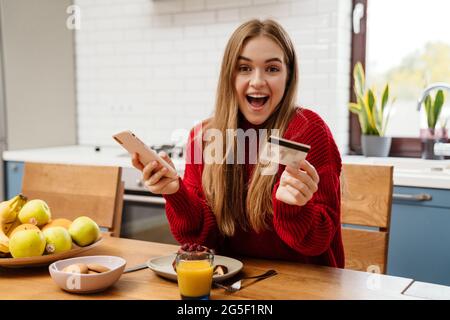 Ziemlich fröhliche junge Frau beim Frühstück, während sie am Küchentisch sitzt, mit Handy und Kreditkarte zum Online-Shopping Stockfoto