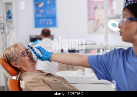 Zahnarzthelferin mit digitaler Körpertemperaturanzeige vor dem auf dem Stuhl sitzenden Patienten. Medizinische Facharzt in der Zahnklinik unter Patienten Temperatur mit digitalen Gerät. Stockfoto