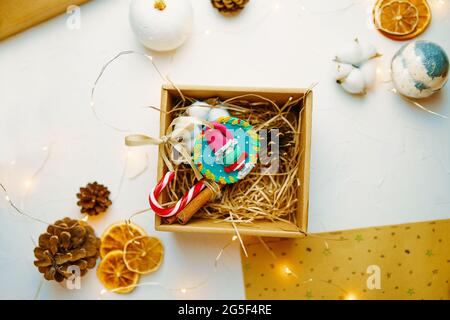 Weihnachts-Geschenkbox mit Souvenir aus Fimo. Niedliche Komposition für die Gestaltung von Grußkarten. Zapfen, getrocknete Orangen, Girlande und Bastelpapier auf weißem Tisch. Neujahrsstimmung. Stockfoto