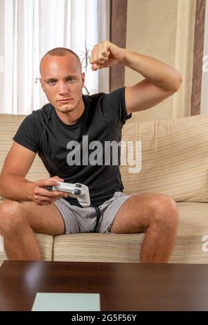 Attraktiver junger Mann mit blauen Augen, der vor Wut die Faust hebt, während er zu Hause eine Spielkonsole im Wohnzimmer spielt. Spielkonzept Stockfoto